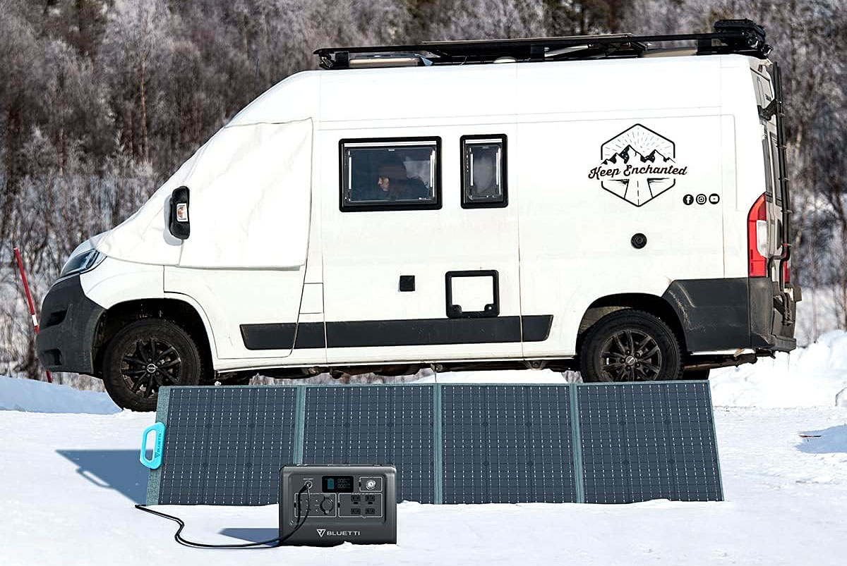 A Bluetti EB70S power station sitting on the snow in front of some solar panels and a camper van