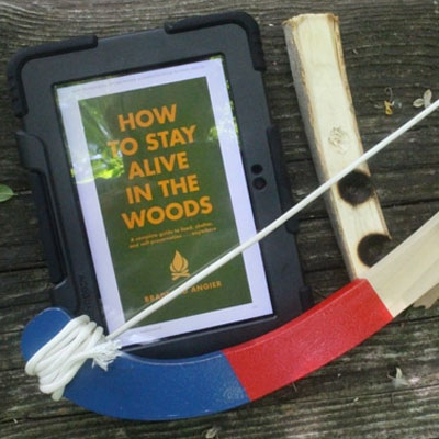 How to stay alive in the woods book on a kindle next to a firemaking bow