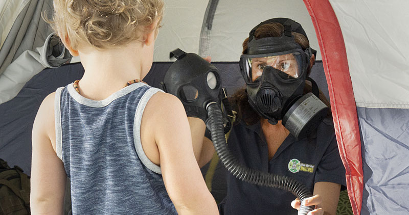 A mom presenting a MIRA Safety Children's Gas Mask CM-2M to her child while sitting in a tent wearing an Avon gas mask