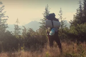 Man wearing a backpack, standing on a hill in the woods, looking over the horizon